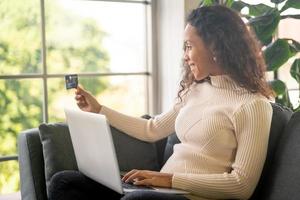 mulher latina usando laptop e segurando um cartão de crédito para fazer compras no sofá foto