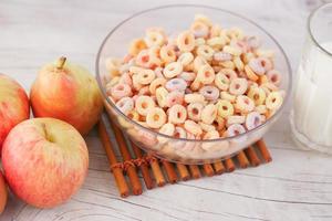 flocos de milho de cereais coloridos em uma tigela, maçã e leite na mesa foto
