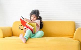garotinha lendo livro com lupa no sofá, crianças felizes brincando com lupa na sala de estar foto