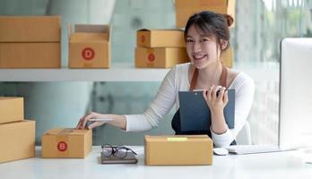 retrato jovem ásia proprietário feminino startup olhar para o trabalho de câmera feliz com caixa em casa preparar entrega de encomendas na cadeia de suprimentos sme, compras, conceito on-line de comércio eletrônico omnichannel. foto