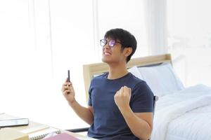 um homem asiático está trabalhando no quarto, ele de bom grado com um smartphone na mão depois de receber as boas notícias. foto