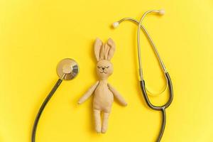 coelho de brinquedo de design simplesmente minimalista e estetoscópio de equipamentos de medicina isolado em fundo amarelo. conceito de médico de crianças de cuidados de saúde. símbolo do pediatra. layout de vista superior plana, copie o espaço foto