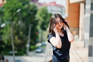 menina morena de vestido preto, óculos de sol posando na rua da cidade. foto
