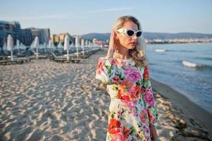 mulher loira de vestido e óculos de sol em pé na praia de areia à beira-mar, apreciando a vista do pôr do sol. foto