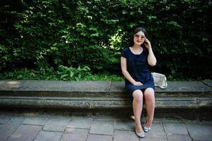 menina de negócios morena de vestido preto em óculos de sol, sentado na fronteira contra arbustos, posando na rua da cidade. foto