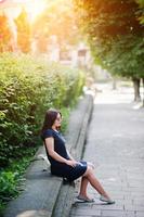 menina de negócios morena de vestido preto em óculos de sol, sentado na fronteira contra arbustos, posando na rua da cidade. foto