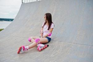 retrato de uma linda garota sentada na pista de patins ao ar livre em shorts, camiseta, óculos de sol e patins. foto