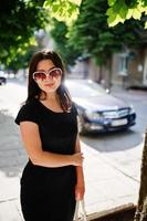 menina de negócios morena de vestido preto em óculos de sol com bolsa na mão posando na rua da cidade, carro de fundo na estrada. foto