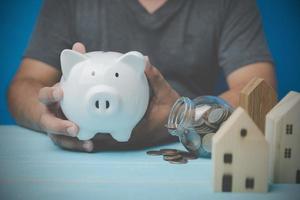 um homem segurando um cofrinho de porco branco na mão com madeira modelo em casa e moedas no vidro. conceito de negociação de casas. habitação, empréstimos bancários para comprar uma casa, investimento imobiliário, negócios imobiliários. foto