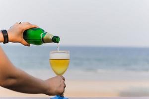 mão de mulher derramando cerveja em vidro na varanda com vista para o mar de férias. foto