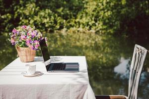 laptop com café e flores na mesa perto da água na zona rural. foto