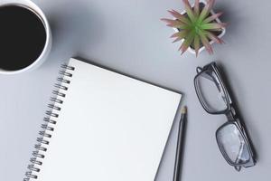 bloco de notas com xícara de café, óculos, caneta e vaso de plantas em uma mesa. postura plana foto