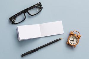 bloco de notas com óculos de leitura, caneta e despertador em fundo azul foto