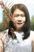 linda mulher asiática de vestido branco sorrindo e mão segurando galhos secos no parque natural. menina tailandesa ou menina chinesa desfrutar de férias com luz solar no jardim foto