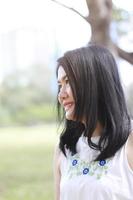 linda mulher asiática de vestido branco relaxa e sorrindo no parque natural. menina tailandesa ou menina chinesa desfrutar de férias com luz solar no jardim foto