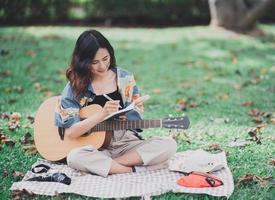 jovem asiática compor ou escrever música e tocar violão no parque, mulher asiática escrevendo música com notebook e tocar violão sentado no gramado. foto