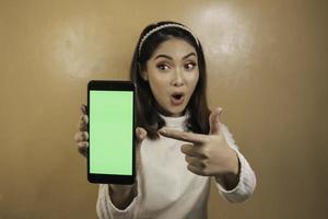 menina bonita asiática está chocada mostra tela verde em smartphone com camisa branca foto