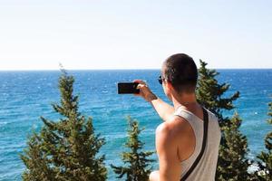 turista masculino tira fotos em um smartphone da bela vista do mar em uma área montanhosa do topo entre árvores coníferas. comunicação de vídeo, usando o telefone ao viajar. cartão sim em roaming