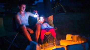 pai e filha sentam-se à noite junto à lareira ao ar livre no verão na natureza. acampamento em família, reuniões ao redor da fogueira. dia dos pais, churrasco. lanterna e barraca de acampamento foto