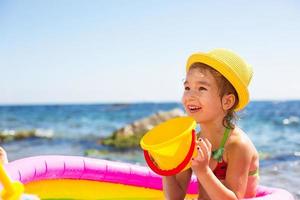 garota de chapéu de palha amarelo brinca com o vento, água e um dispensador de água em uma piscina inflável na praia. produtos indeléveis para proteger a pele das crianças do sol, queimaduras solares. resort no mar. foto