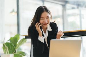 feliz jovem asiática falando no celular e sorrindo enquanto está sentado em seu local de trabalho no escritório. foto