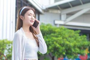 jovem profissional de negócios asiáticos de vestido branco está ligando para o telefone seriamente com funcionários de escritório no prédio ao ar livre e arbustos como pano de fundo. foto