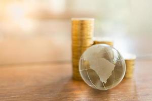 negócios globais e conceito financeiro. close-up da mini bola do mundo de cristal claro com pilha de moedas de ouro na mesa de madeira e copie o espaço. foto