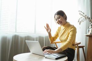 mulher asiática tendo chamada de vídeo em seu computador em casa. garota sorridente estudando online com o professor.