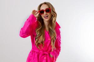 Feche a garota feliz em óculos de sol vermelhos e casaco rosa isolado no fundo branco. jovem animado, chocado e surpreso com roupas da moda. conceito de moda e compras. longos cabelos claros e ondulados. foto