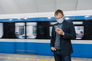 passageiro masculino usa máscara facial posa na plataforma, espera o trem, viaja de metrô, concentrado no dispositivo smartphone, lê notícias online. consciência do vírus em local público. surto de coronavírus foto