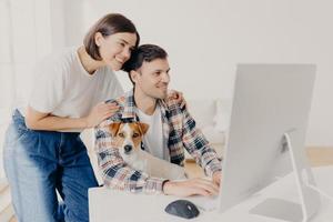 imagem de mulher toca suavemente os ombros do marido, olha positivamente no monitor do computador, navega na internet, compra móveis on-line para seu novo apartamento. homem feliz trabalha no dispositivo moderno com cachorro foto