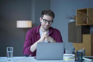 funcionário de home office pensativo jovem alemão senta-se na mesa no local de trabalho opera on-line no laptop foto