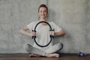 mulher alegre fitness com roupa ativa, sentado em posição de lótus e se exercitando com anel de pilates foto