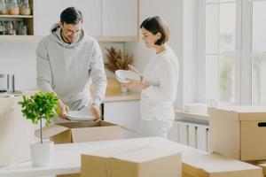 foto de mulher casada feliz e homem desempacotar louça de caixas, vestida casualmente, segurar pratos, posar na nova cozinha moderna, cercada de pacotes em movimento. conceito de pessoas, casa e realocação