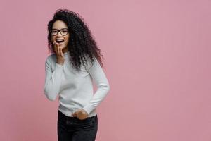 mulher afro-americana positiva ri de algo engraçado, cobre a boca aberta com a palma da mão, não pode controlar suas emoções, mantém a mão no bolso da calça jeans, usa poloneck branco, isolado no fundo rosa foto