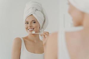 mulher jovem e bonita com sorriso perfeito saudável escovando os dentes e olhando no espelho foto