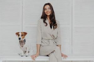 imagem de mulher europeia morena atraente senta-se junto com o cão de pedigree, vestido com roupas da moda, olha com um sorriso gentil para a câmera. pessoas, cuidados com os animais e conceito de responsabilidade foto