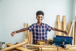 carpinteiro de menino afro-americano sorridente, abrindo os braços alegremente para mostrar que a madeira é colocada na mesa tentará ter sucesso foto