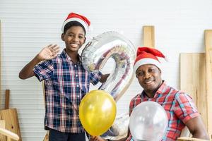 natal feliz filho e pai. alegre carpinteiro de filho afro-americano em chapéus de Papai Noel se divertindo sorrindo para a câmera acenando e dizendo olá para você com o pai, aproveitando o tempo juntos foto