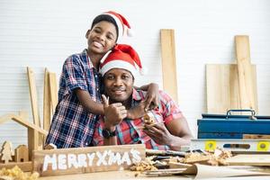 natal feliz filho e pai. alegre carpinteiro de filho afro-americano abraçando seu pai enquanto se inclinava na mesa de madeira com diversas ferramentas de trabalho deitadas sobre ela foto