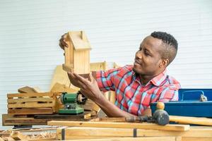 jovem carpinteiro afro-americano olha com orgulho para uma casa de madeira modelo. quando tiver sucesso foto