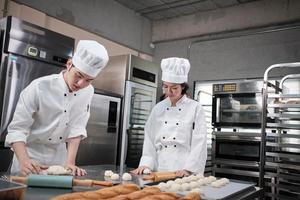 dois chefs asiáticos profissionais em uniformes e aventais de cozinheiro branco amassam massa e ovos, preparam pão, biscoitos e alimentos frescos de padaria, assando em um forno em uma cozinha de aço inoxidável de restaurante. foto