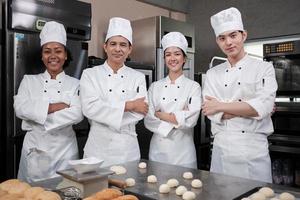 um grupo de funcionários da equipe de chefs felizes profissionais multirraciais em uniformes de cozinha brancos fica em uma fileira, braços cruzados com confiança, sorrisos alegres com trabalhos de culinária comercial na cozinha do restaurante.