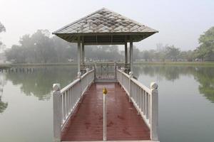 ponte vermelha conecta entre o pavilhão branco em mash e a terra, atrás das árvores e reflete na água no pântano pela manhã, uma pequena cobertura de neblina acima, tailândia. foto