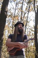 macho barbudo com cabelo comprido e chapéu posando com ukulele na floresta foto