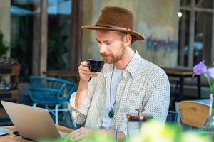 jovem hipster elegante no trabalho de chapéu com laptop ao ar livre no café de rua foto