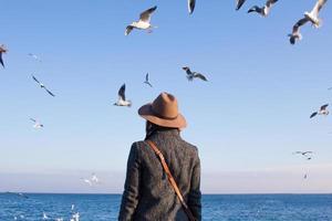 jovem mulher de chapéu na praia de outono n dia ensolarado e assistindo gaivotas foto