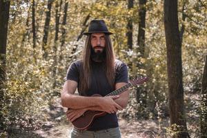 macho barbudo com cabelo comprido e chapéu posando com ukulele na floresta foto