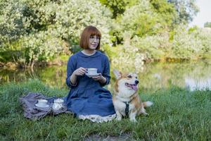 jovem de vestido retrô com cachorro corgi engraçado no piquenique, fêmea com cachorro fofo bebe chá inglês no parque foto