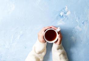 mulher segurando uma xícara de chá quente na mesa azul, mãos no suéter quente com caneca, manhã de inverno foto